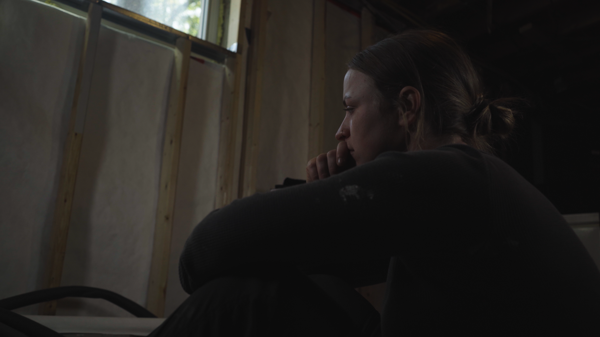 Woman sitting alone in a dimly lit room under construction, staring out a small window deep in thought