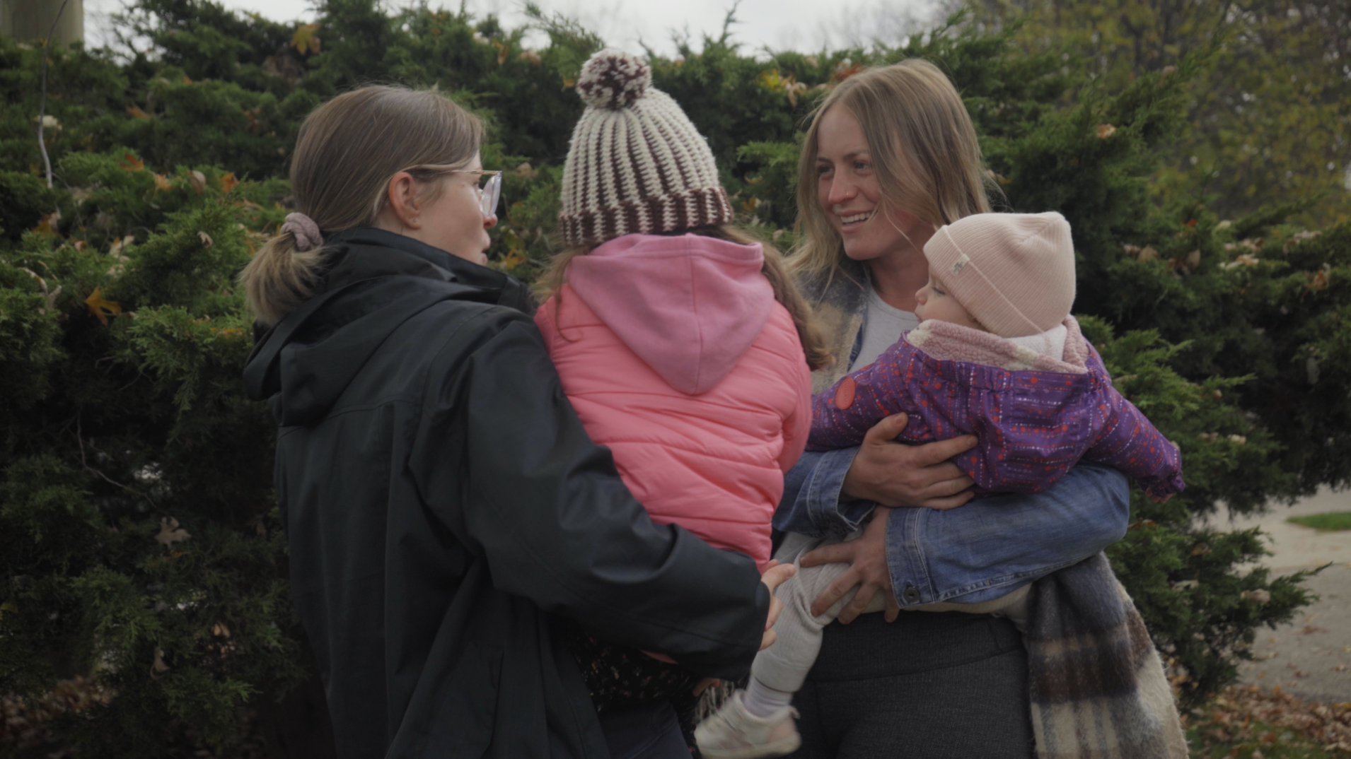 Two women smiling at each other while holding toddlers bundled up in winter coats.