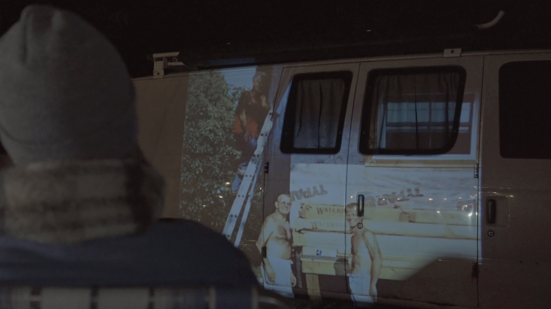 Rachel watching home videos projected onto the side of a van at night