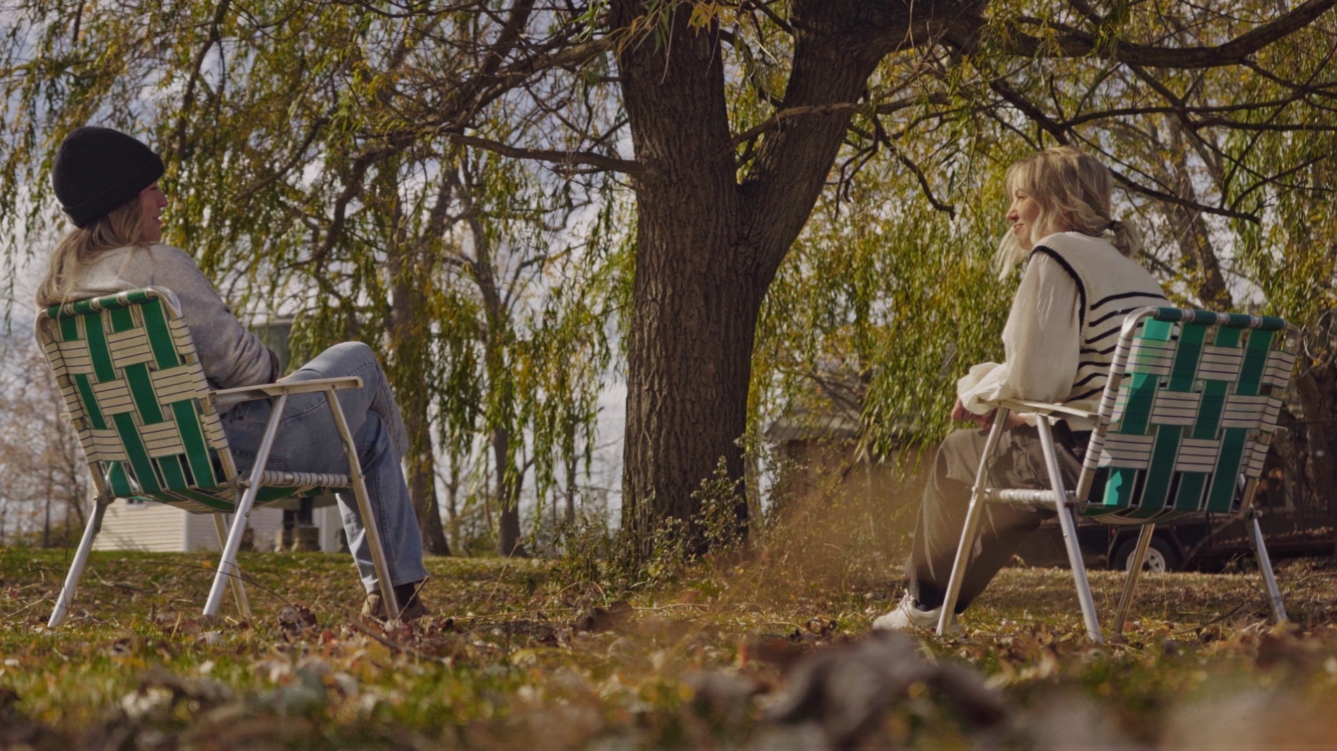 Two women sitting on lawn chairs under a willow tree, having a quiet outdoor conversation